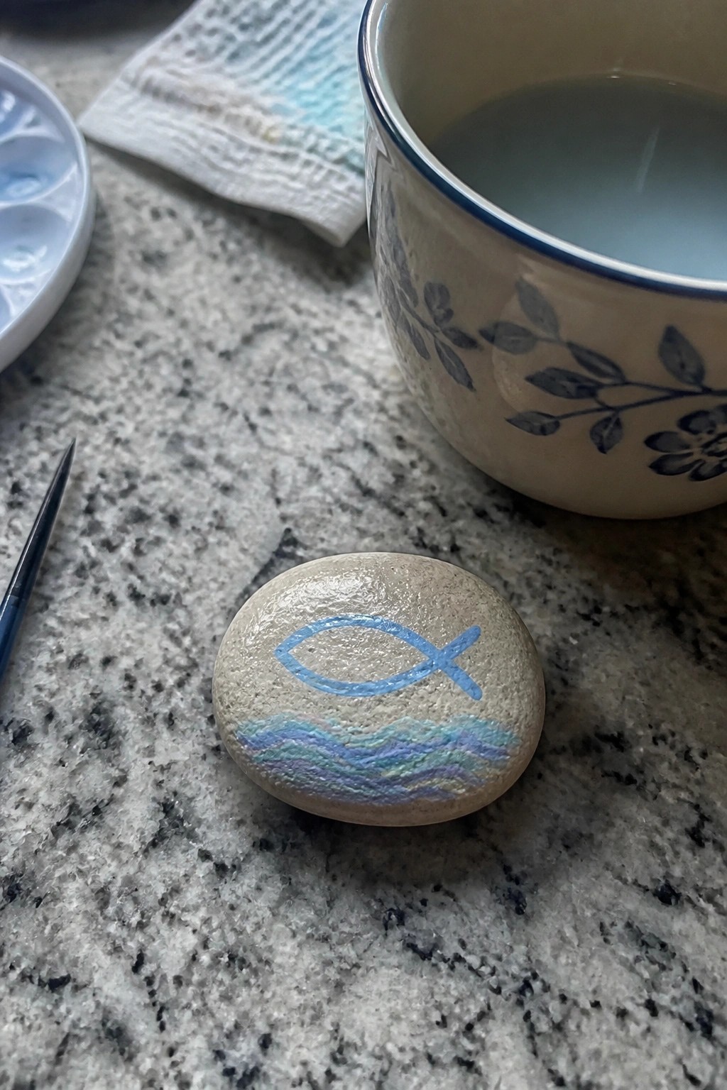 Round stone painted with blue fish symbol and waves beside floral ceramic mug on granite