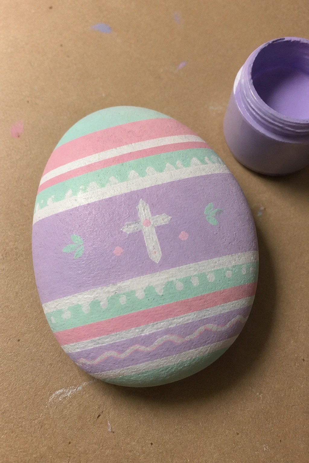 Pastel striped egg with cross design beside open jar of purple paint