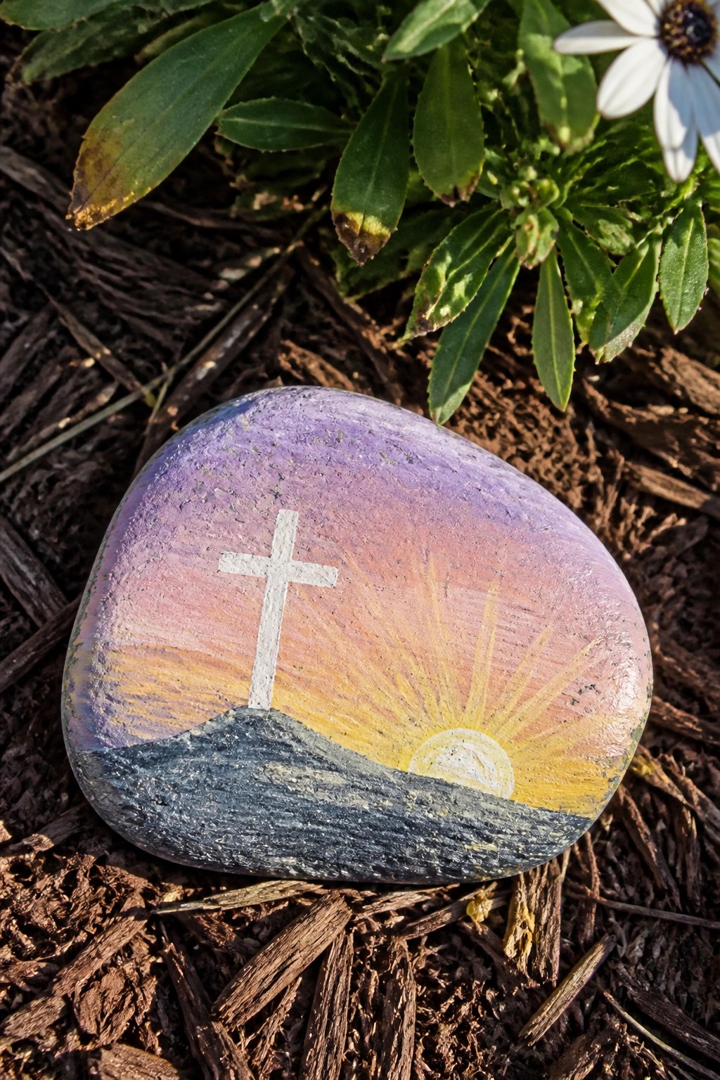 Painted rock with white cross and sunrise over hills amid mulch and plants