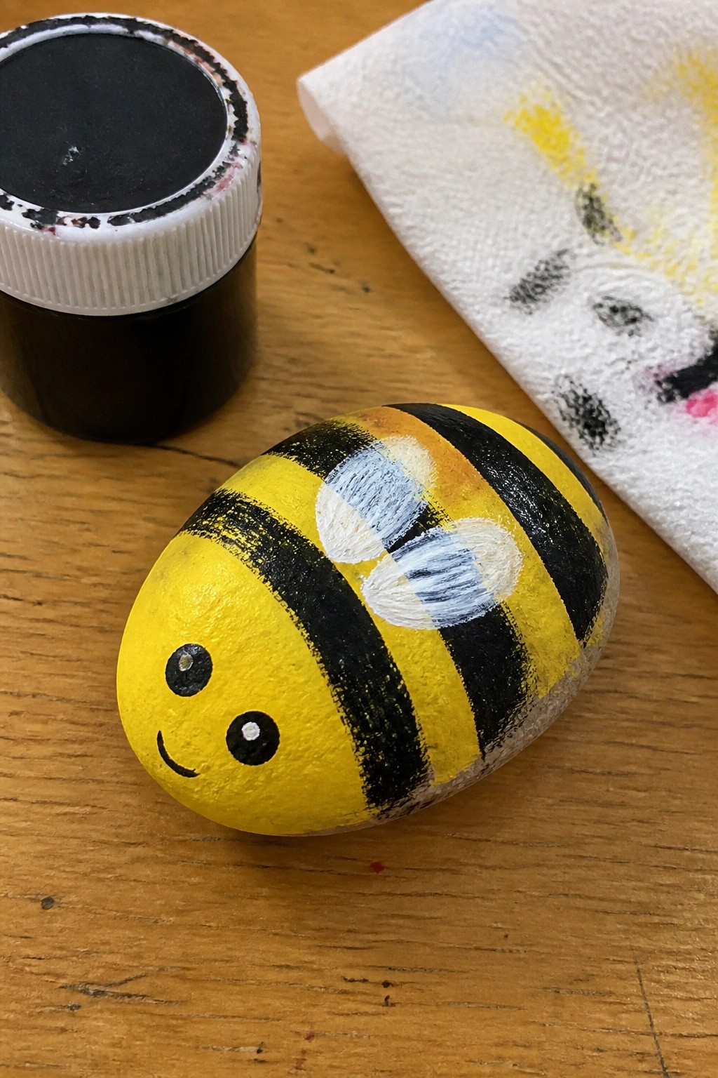 Painted smiling bee rock with yellow-black stripes and wings beside paint jar on wood