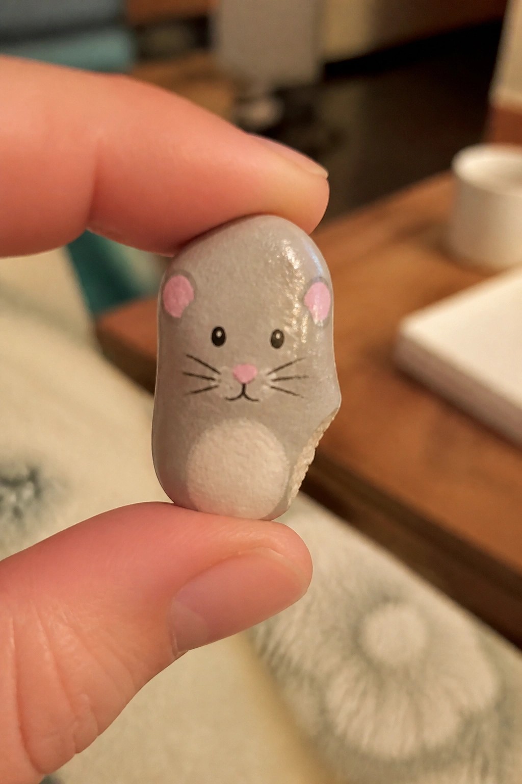 Fingers holding a small gray stone painted as a mouse with pink ears.