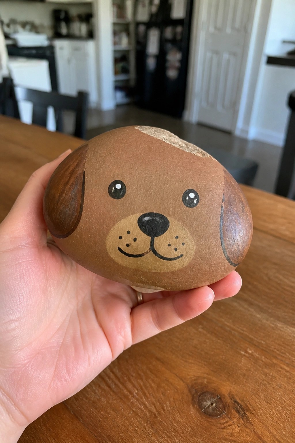 Hand holding a brown rock painted as a dog face on wooden table.