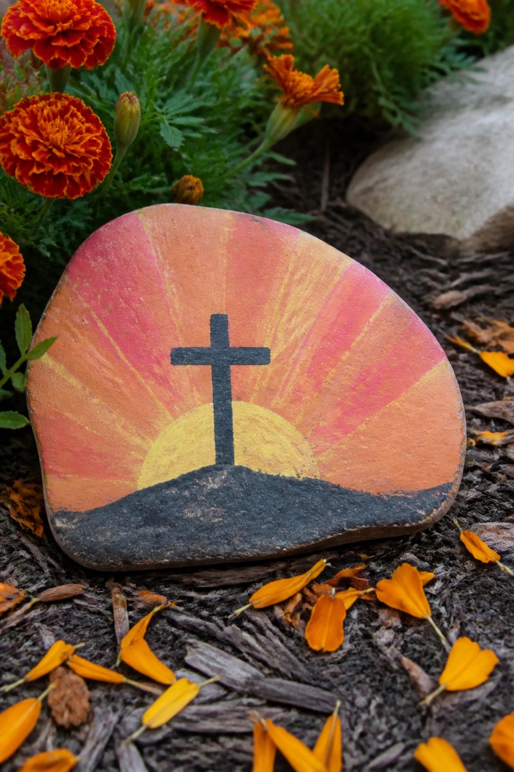 Painted rock with black cross over yellow sun amid orange marigolds and mulch