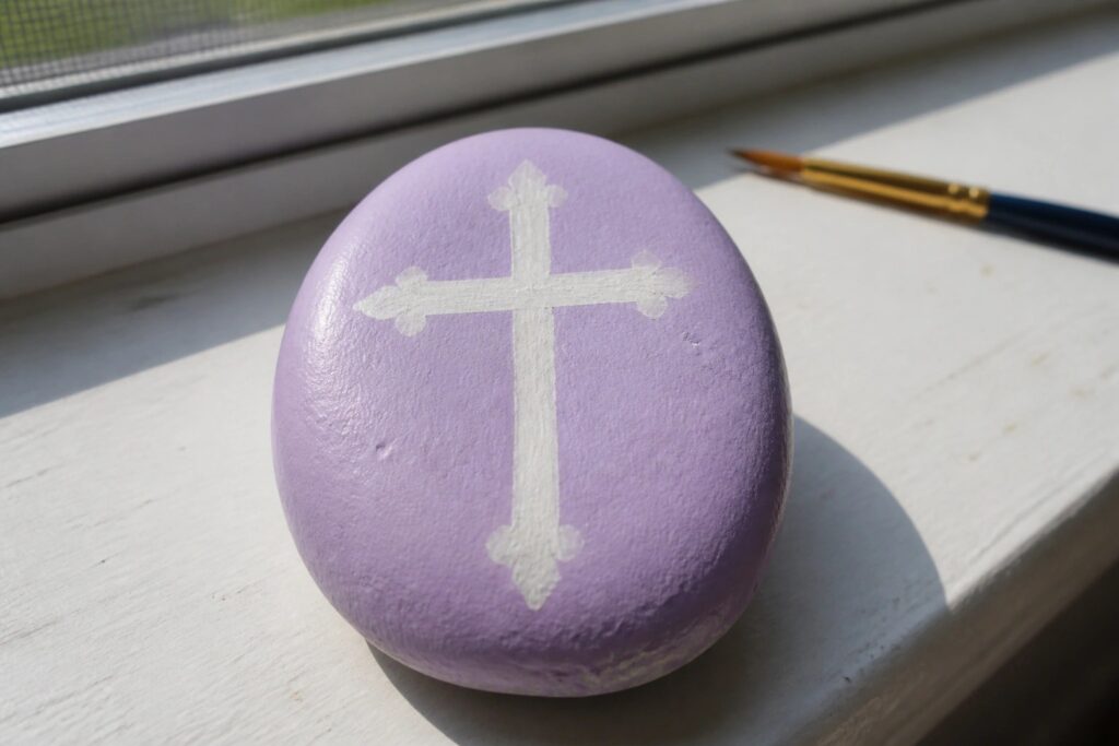 A smooth oval stone painted lavender with a white cross centered on top sits on a white windowsill next to a paintbrush.