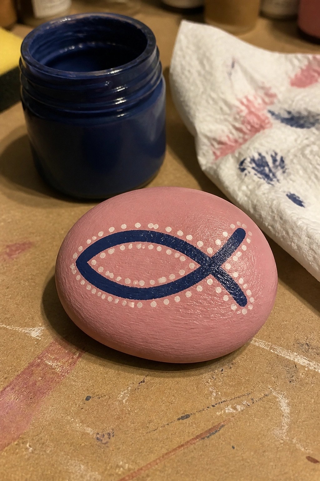 Pink painted rock with blue fish symbol beside open paint jar and stained paper towel