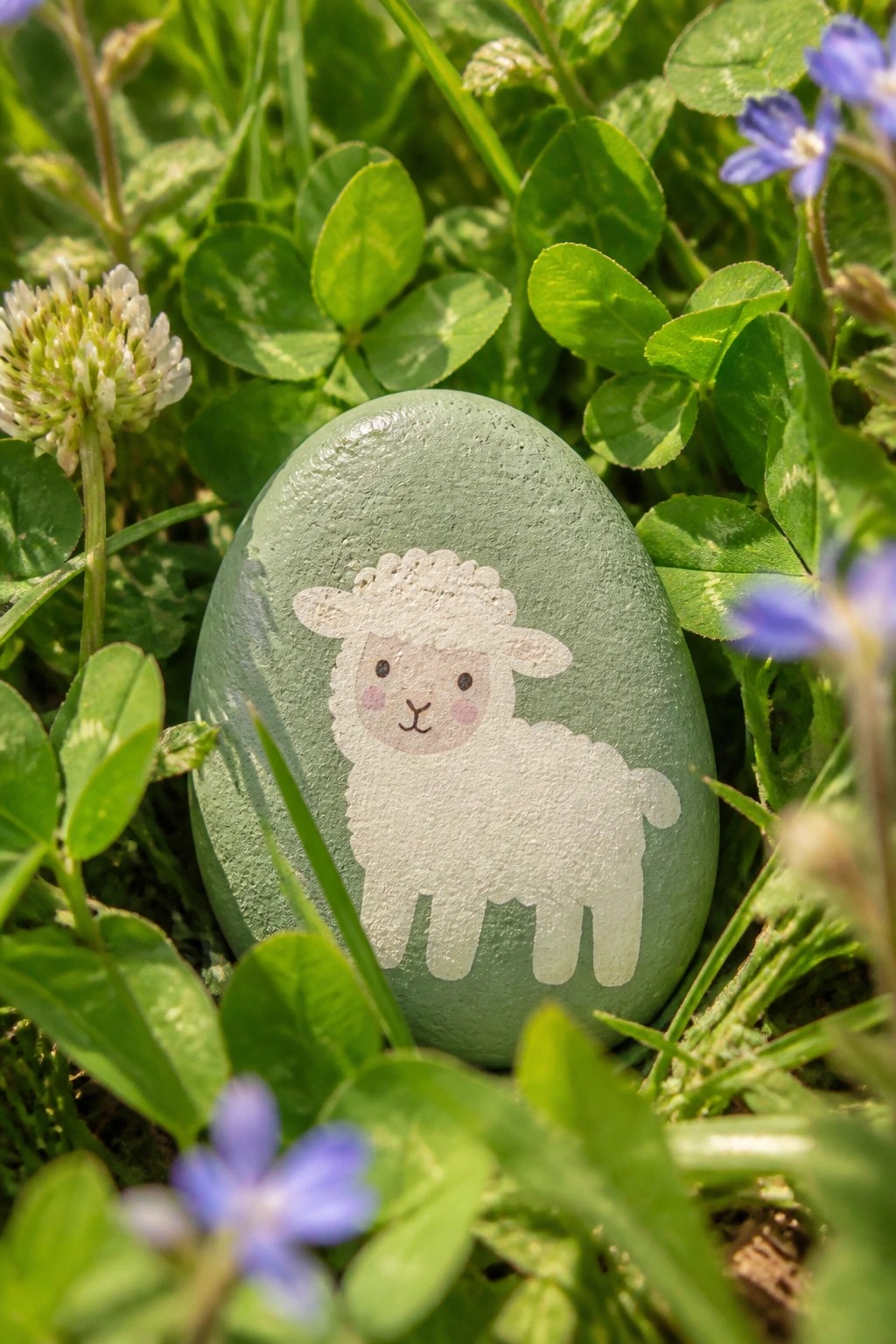 Green painted rock with white sheep design amid clover leaves and blue flowers