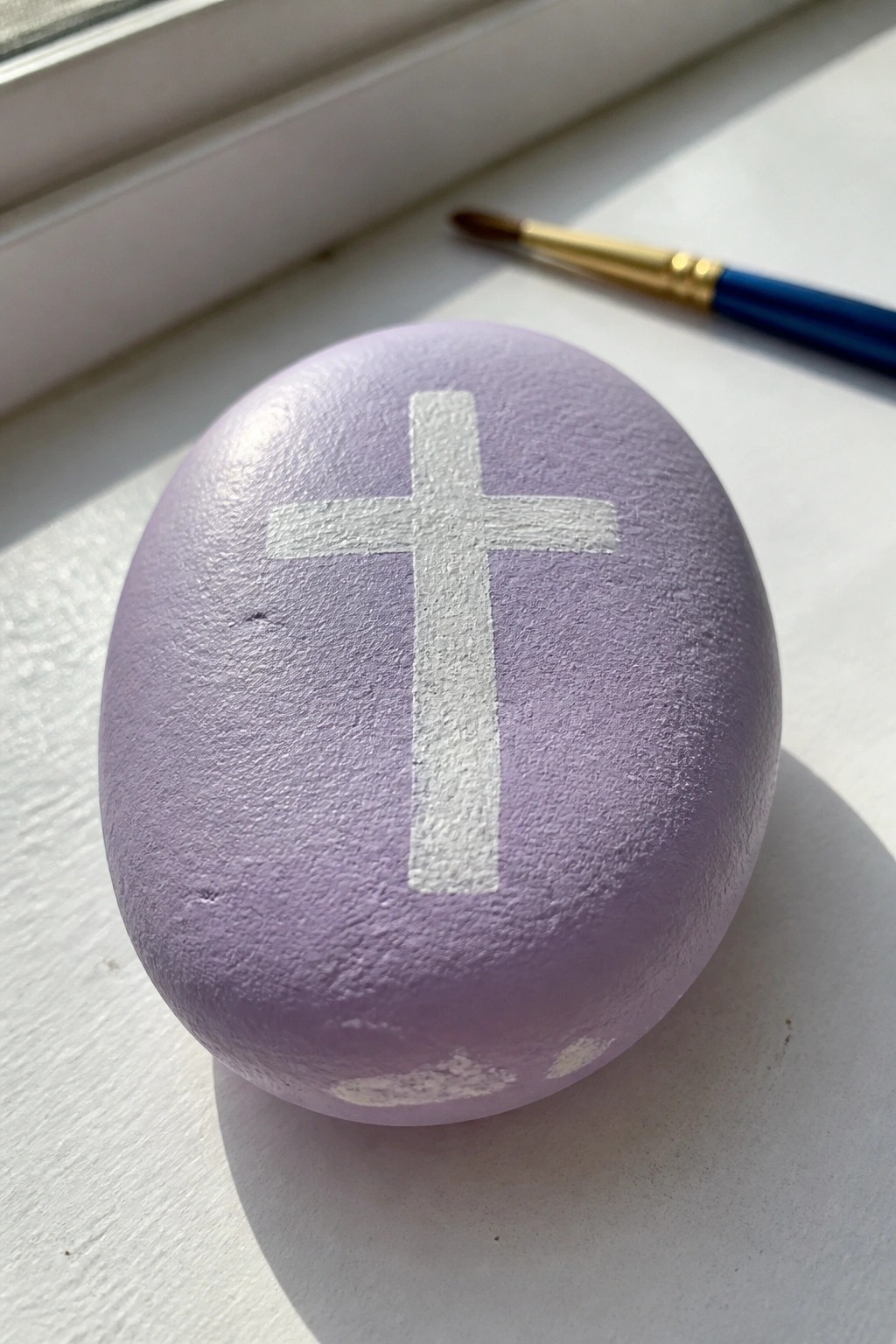 A smooth oval stone painted lavender with a white cross centered on top sits on a white windowsill next to a paintbrush.
