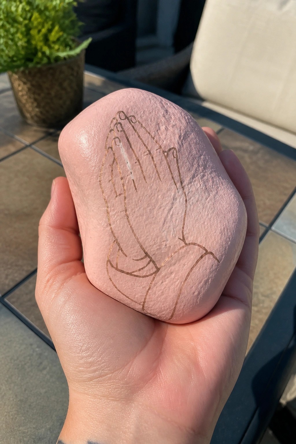 Hand holding pink stone engraved with golden praying hands on patio