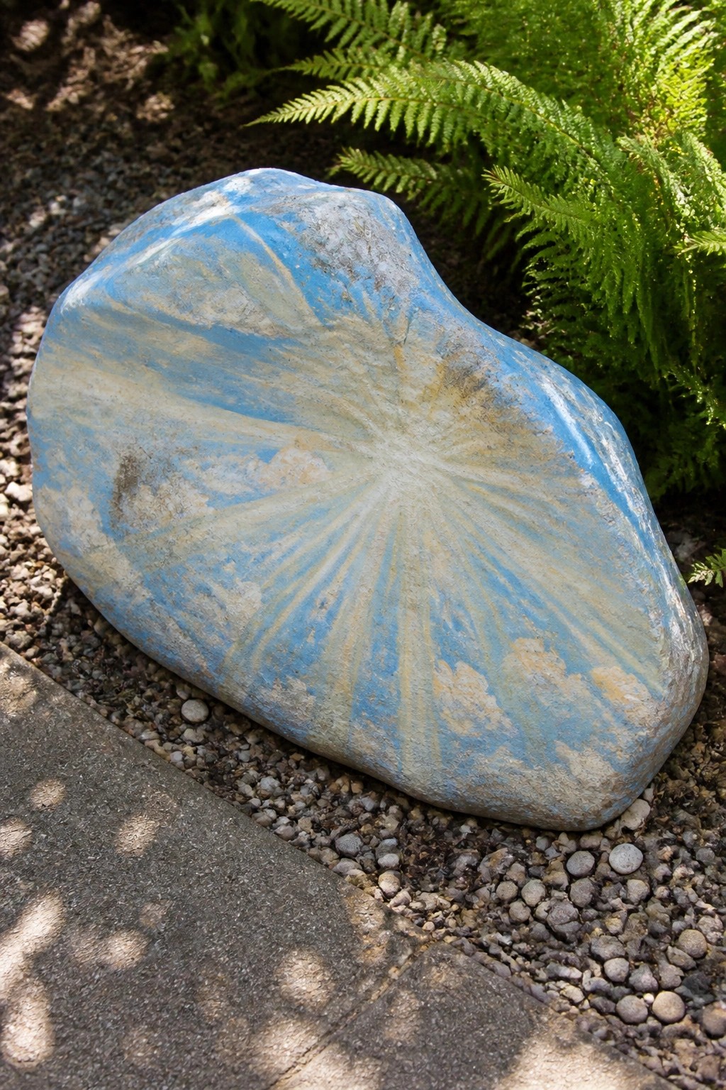 Blue painted rock with radiating white and yellow starburst pattern on gravel.