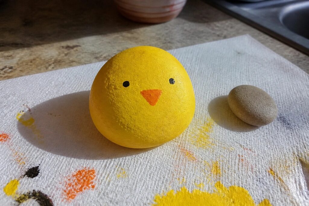 Yellow chick rock with orange beak beside small pebble on paint-splattered white cloth