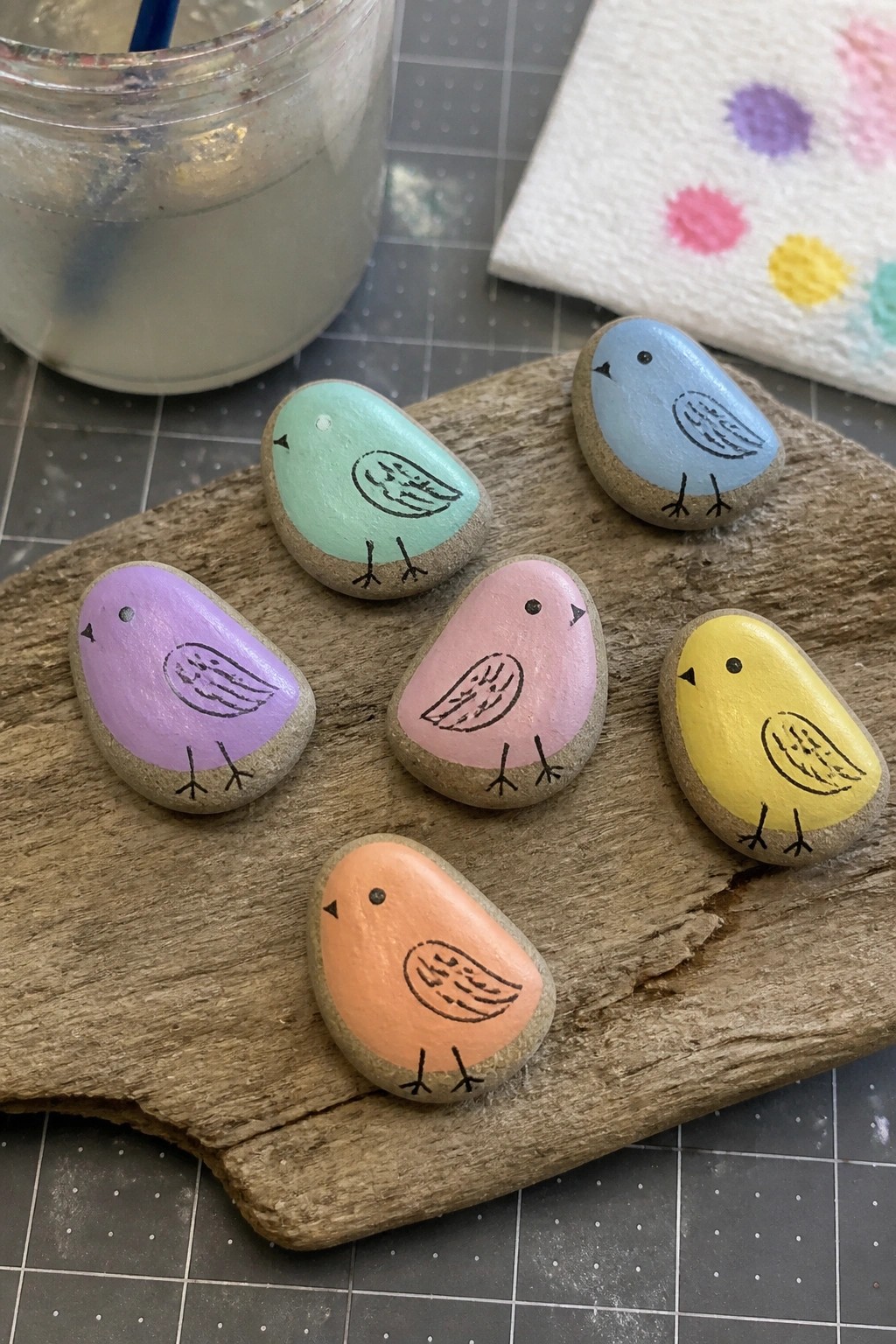 Six pastel bird-shaped rocks painted on stones atop a wooden board