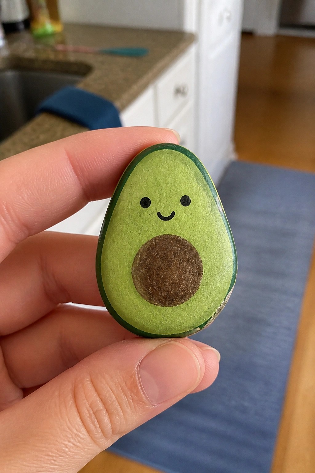 Hand holding smiling avocado painted on green stone with brown pit in kitchen