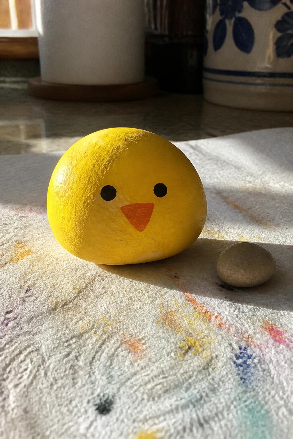 Yellow chick rock with orange beak beside small pebble on paint-splattered white cloth