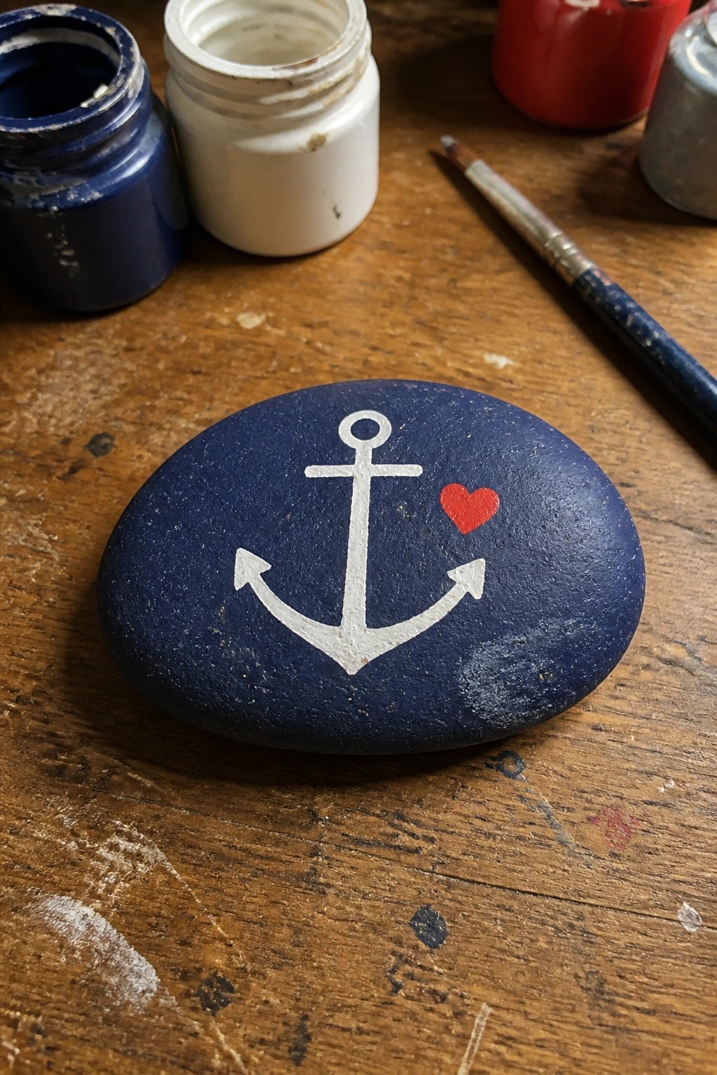 Blue painted rock with white anchor and red heart beside paint jars and brush on wood