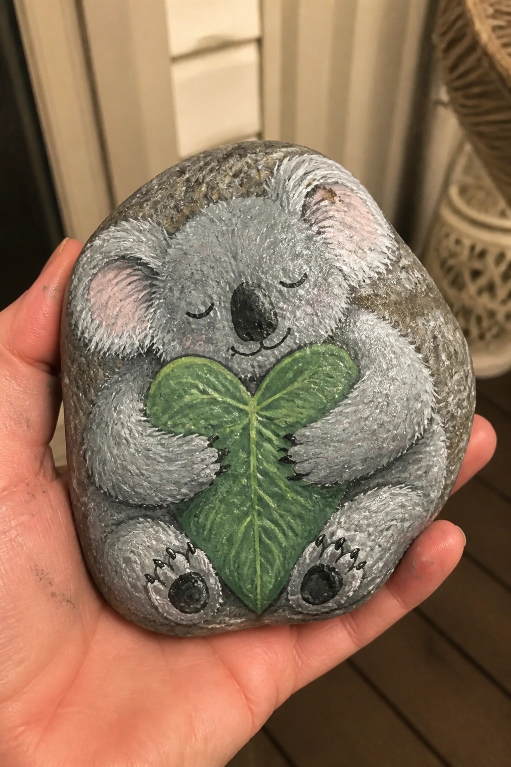 Hand holding painted rock of gray koala hugging green leafy heart.