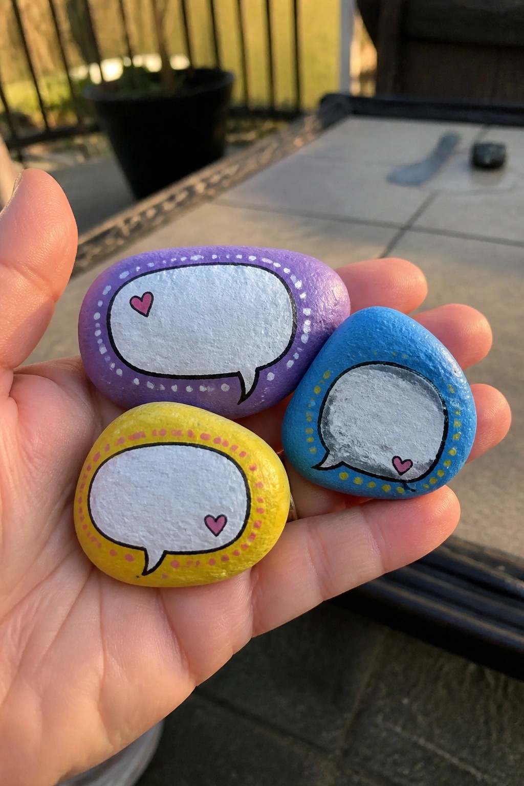 Hand holding three painted rocks resembling speech bubbles in purple, yellow, and blue.