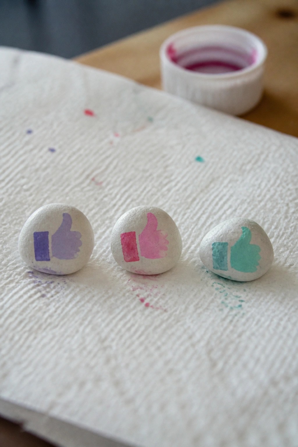 Three white pebbles with purple, pink, and teal thumbs-up icons on paper towel
