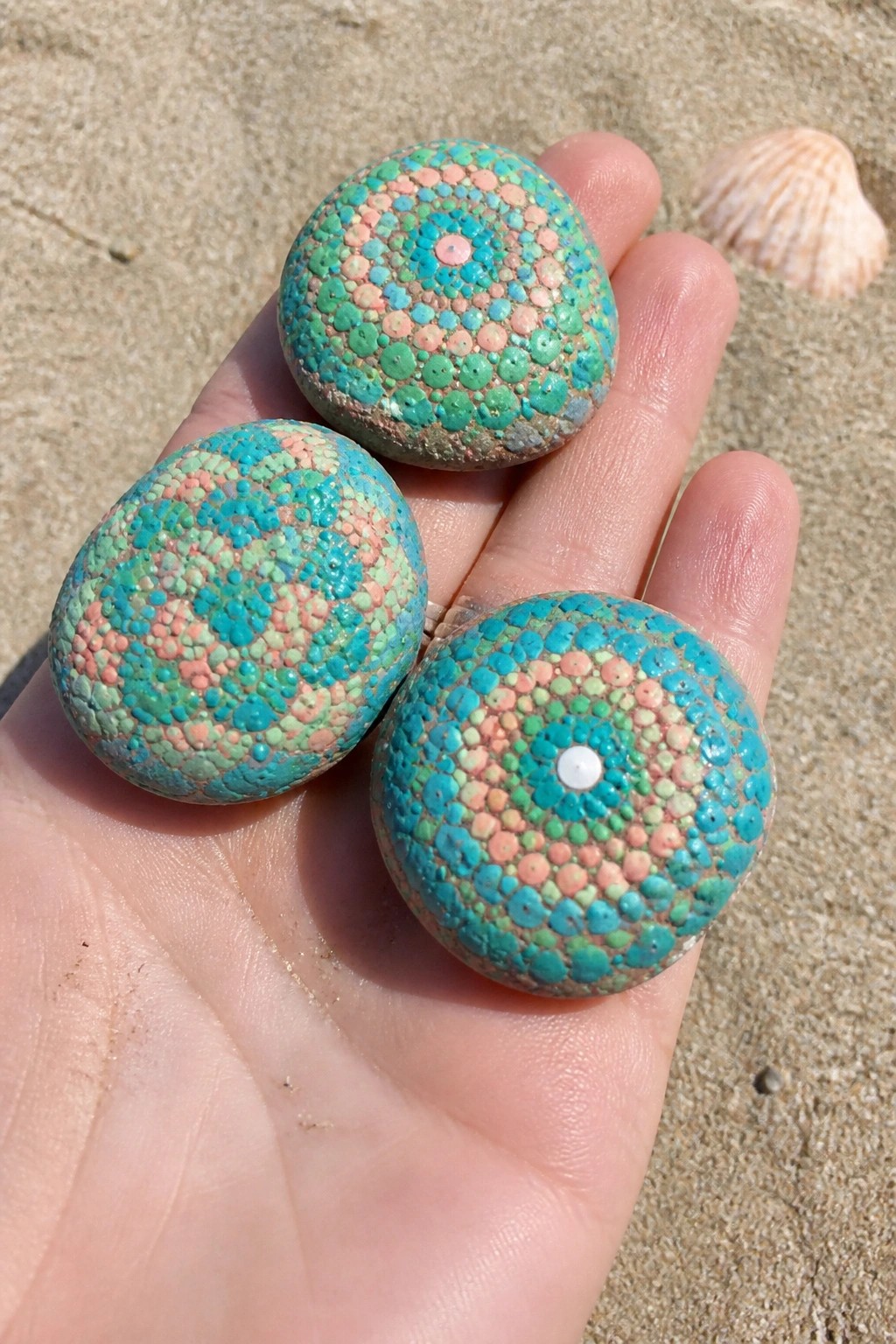 Hand holding three turquoise, pink, and green dotted painted rocks on sand.