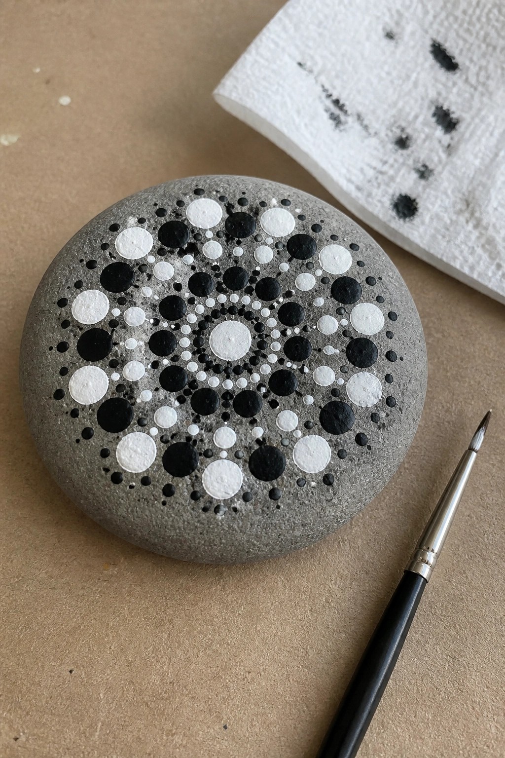 Gray stone with black and white dot mandala, paintbrush and paper towel nearby.