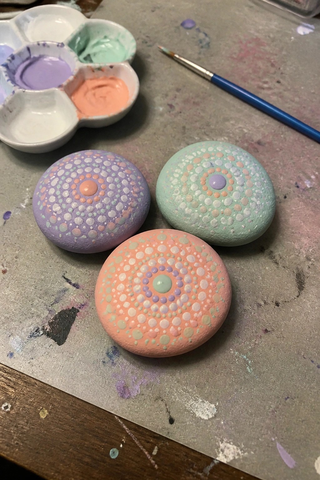 Three pastel dotted rocks beside paint palette and brush on messy table