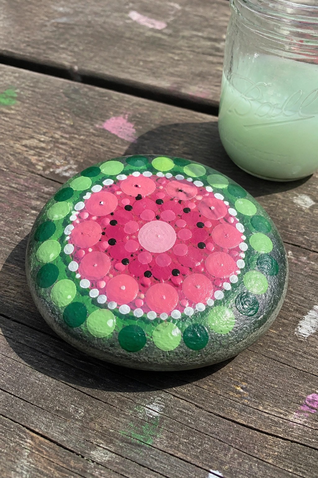 Watermelon dot-painted rock beside glass jar on splattered wooden table