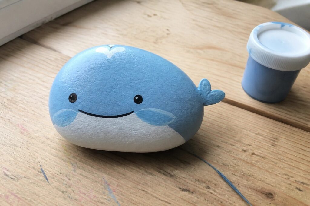 Blue whale painted rock with cute face beside paint jar on wooden table.