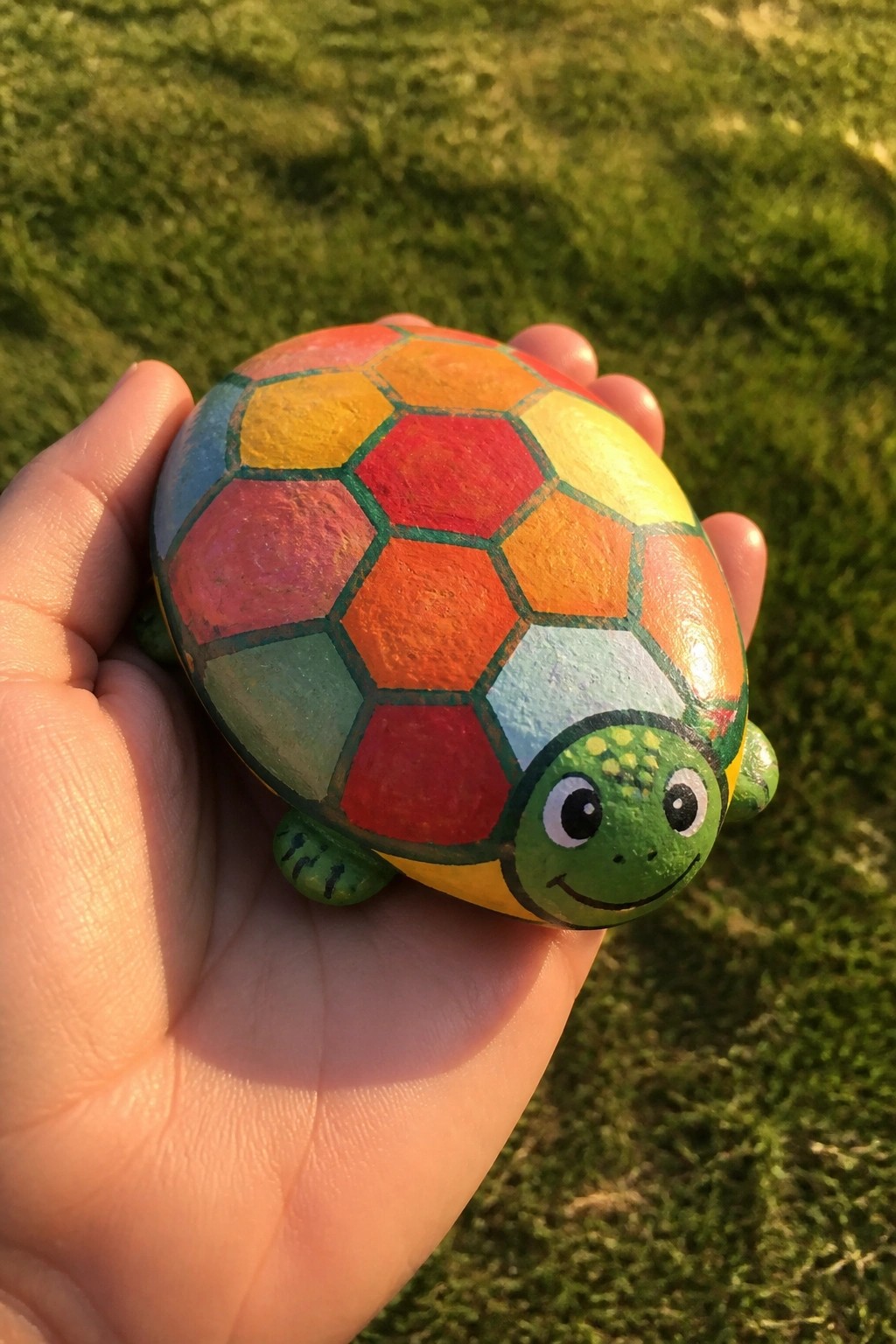 Hand holding rainbow-painted turtle rock with green smiling head on grass.
