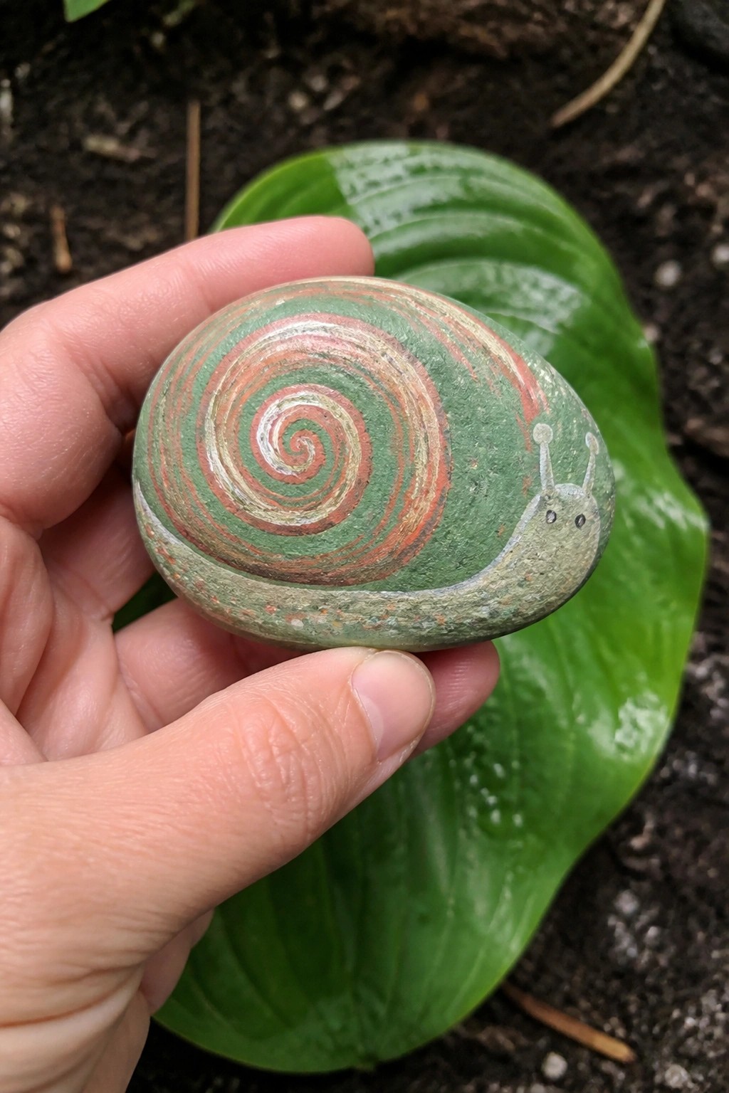 Hand holds painted rock snail with green body and red-white spiral shell over leaf