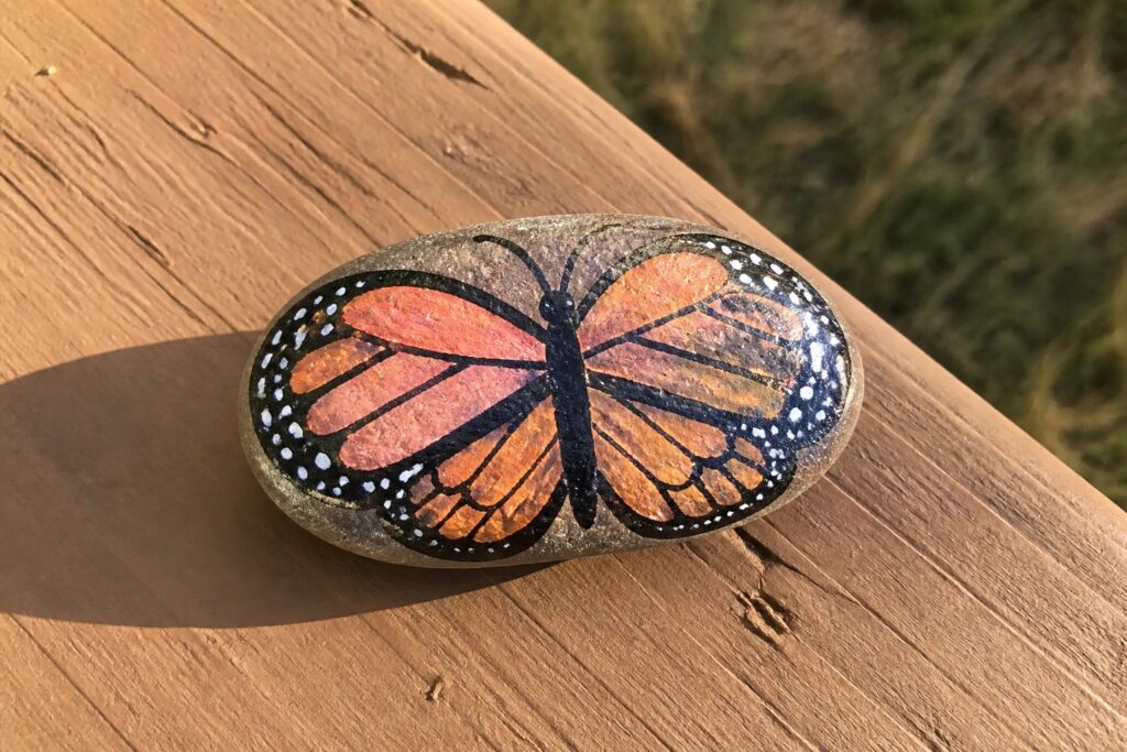 Oval rock painted with orange monarch butterfly and black markings on wood