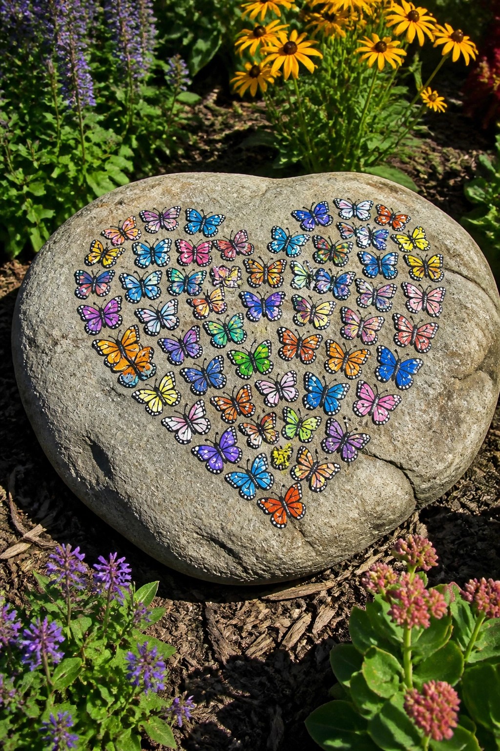 Colorful butterflies arranged in a heart on a large gray garden rock amid flowers