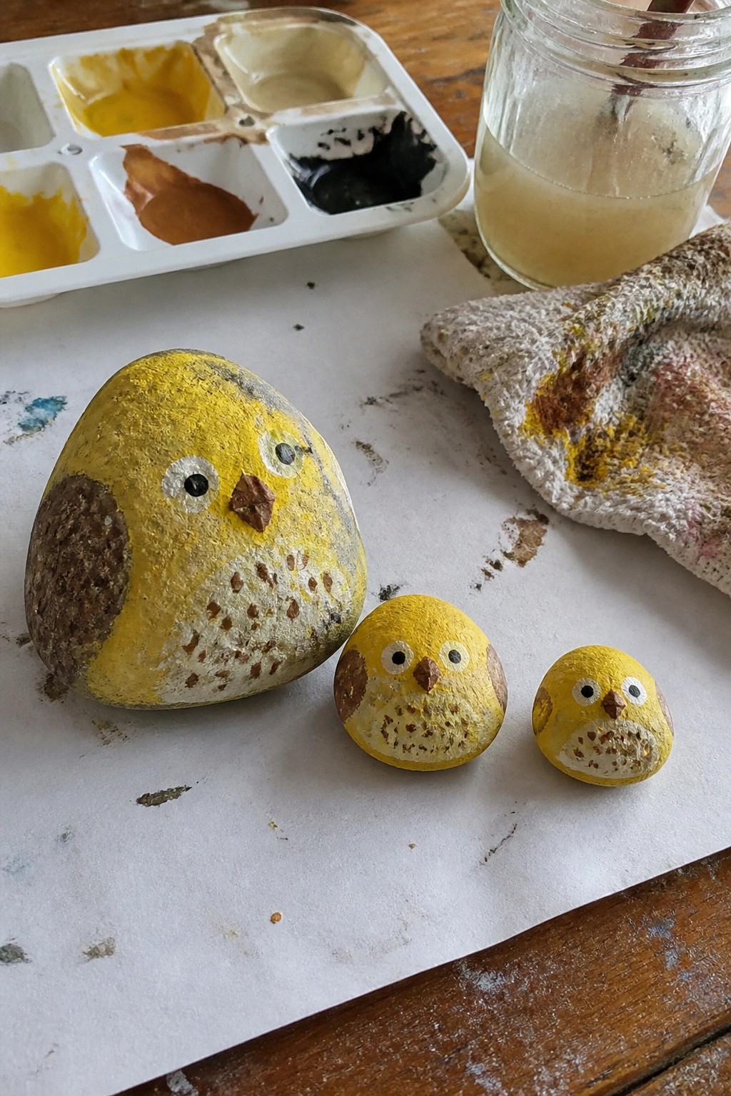 Three yellow painted owl rocks beside palette, jar, and stained cloth