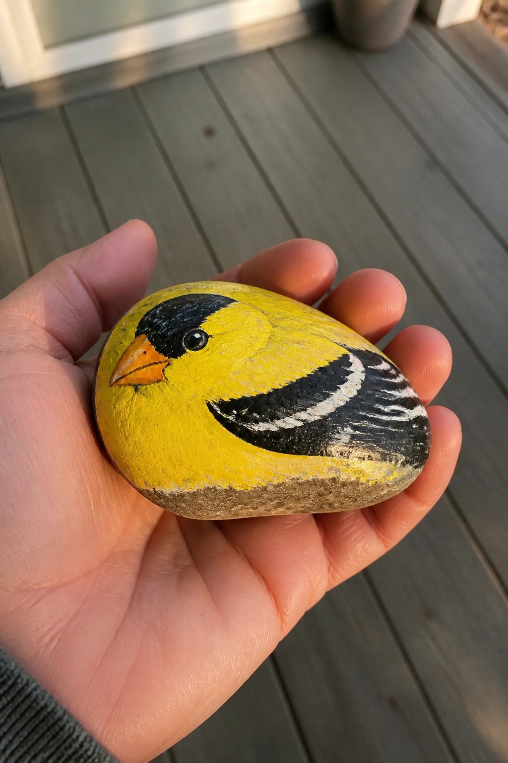 Hand holding yellow stone painted as bird with black head and orange beak.