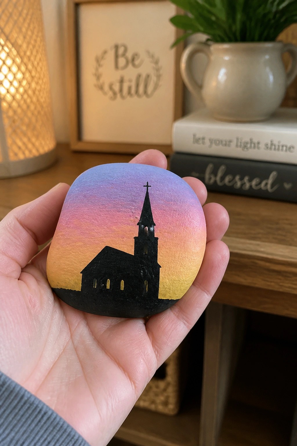 Hand holding painted rock with sunset gradient and black church silhouette.