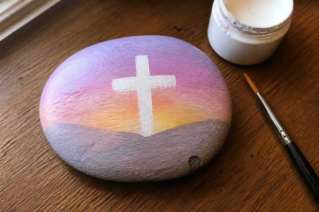 Painted rock with white cross on purple-orange sunset gradient beside paint jar and brush