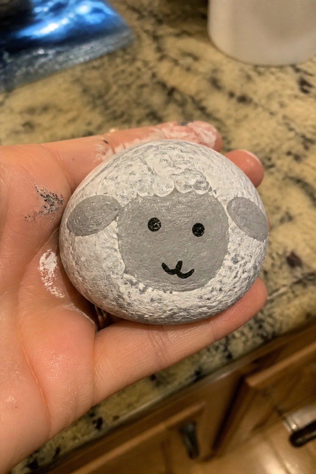 Hand holding round stone painted as sheep with gray face and ears.