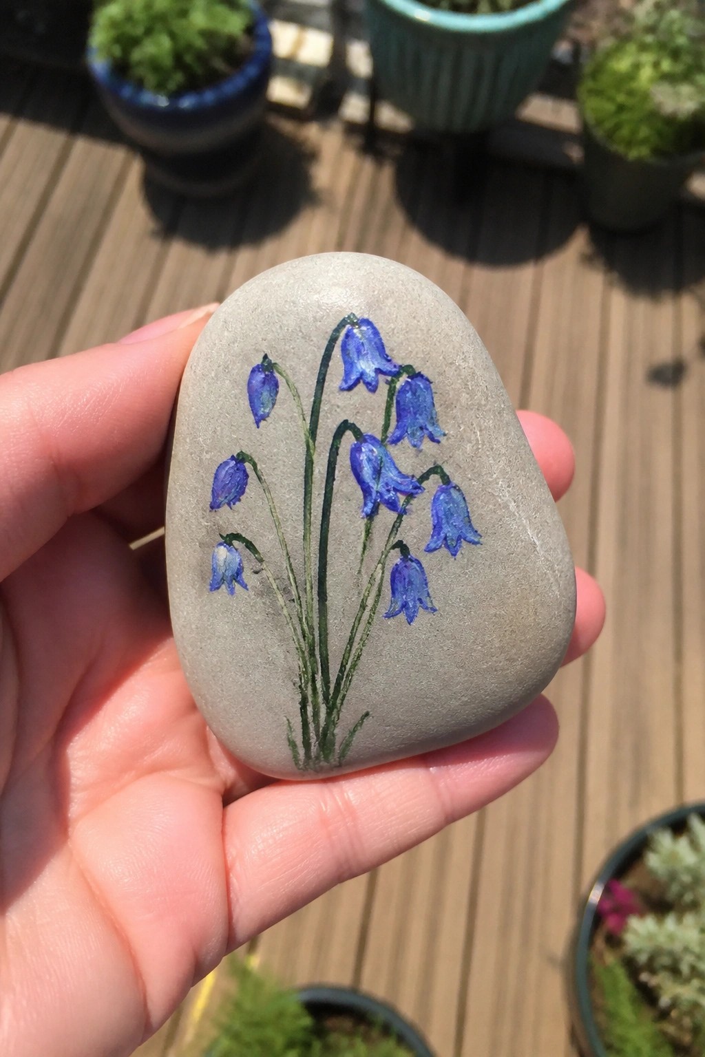A hand holds a smooth gray oval rock painted with blue bell-shaped flowers on green stems.