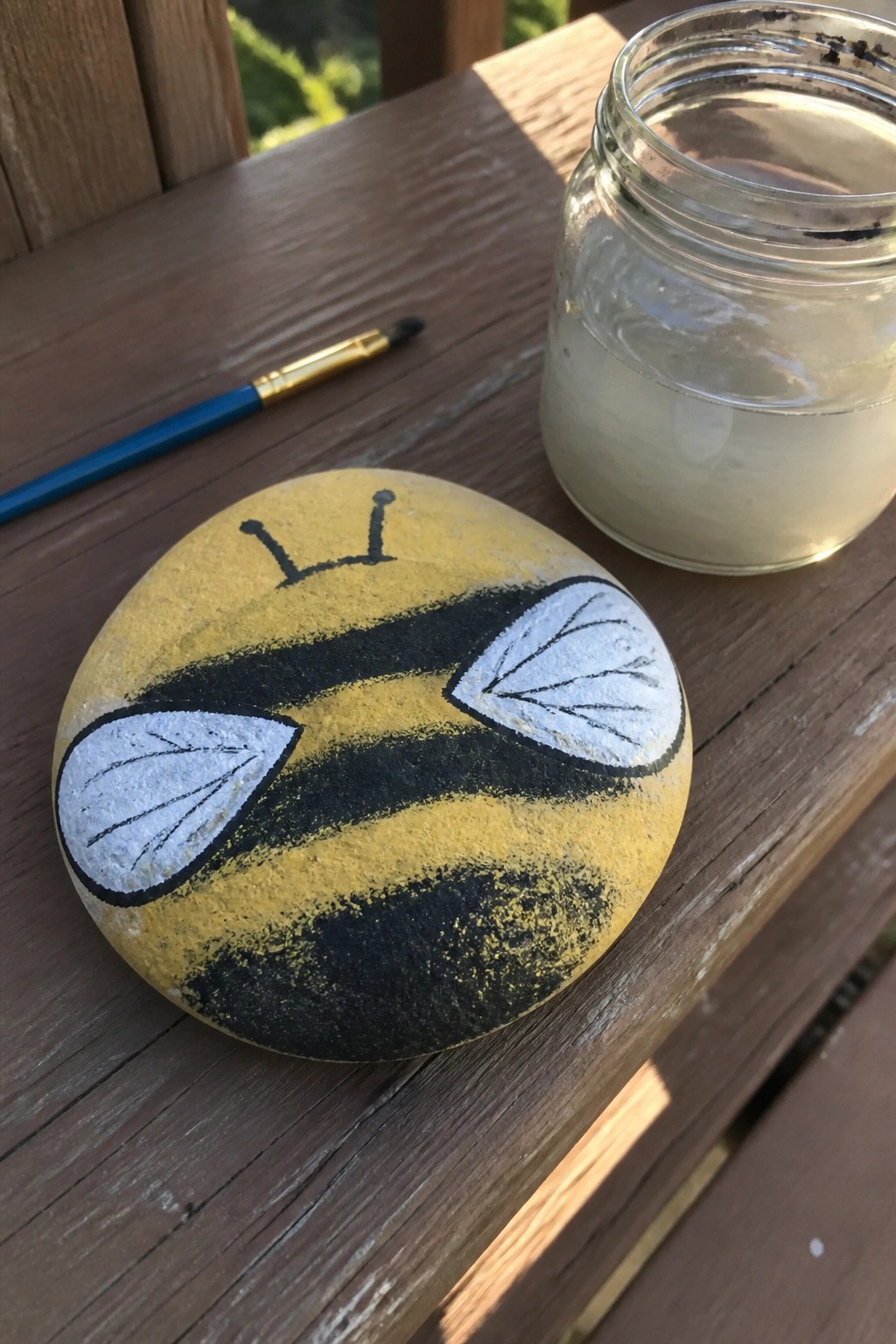Yellow bumblebee painted rock beside jar and brush on wooden table