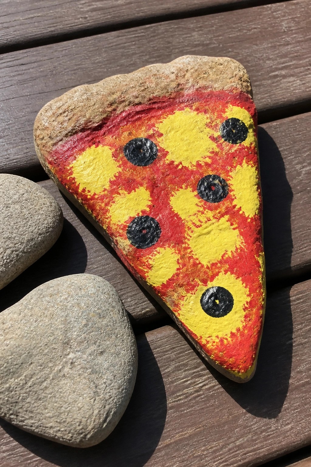 Painted rock shaped as pizza slice with red sauce, yellow cheese, black olives on wood