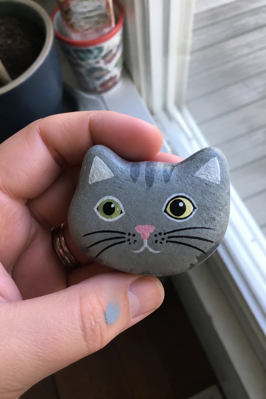 Hand holding gray rock painted as cat with green and yellow eyes