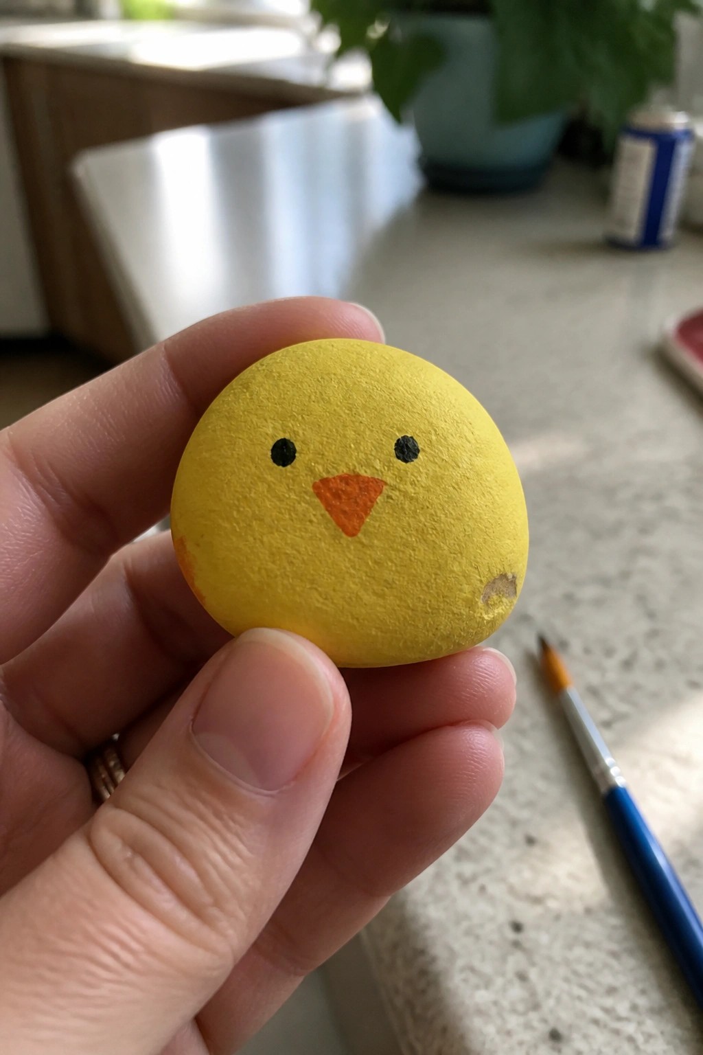 Hand holding yellow painted rock chick with black eyes and orange beak.