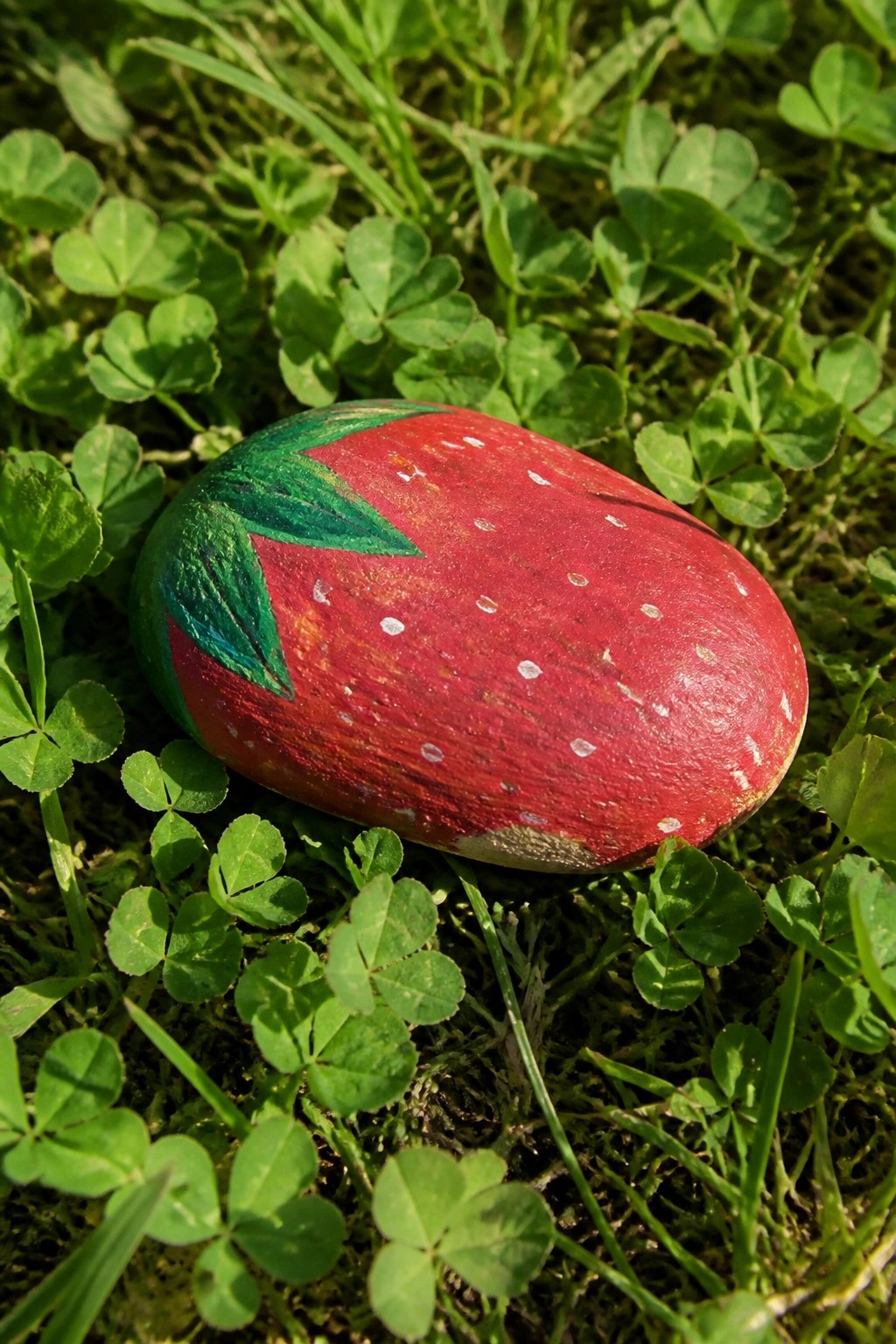 Red strawberry-painted rock with white seeds and green leaves on clover grass.