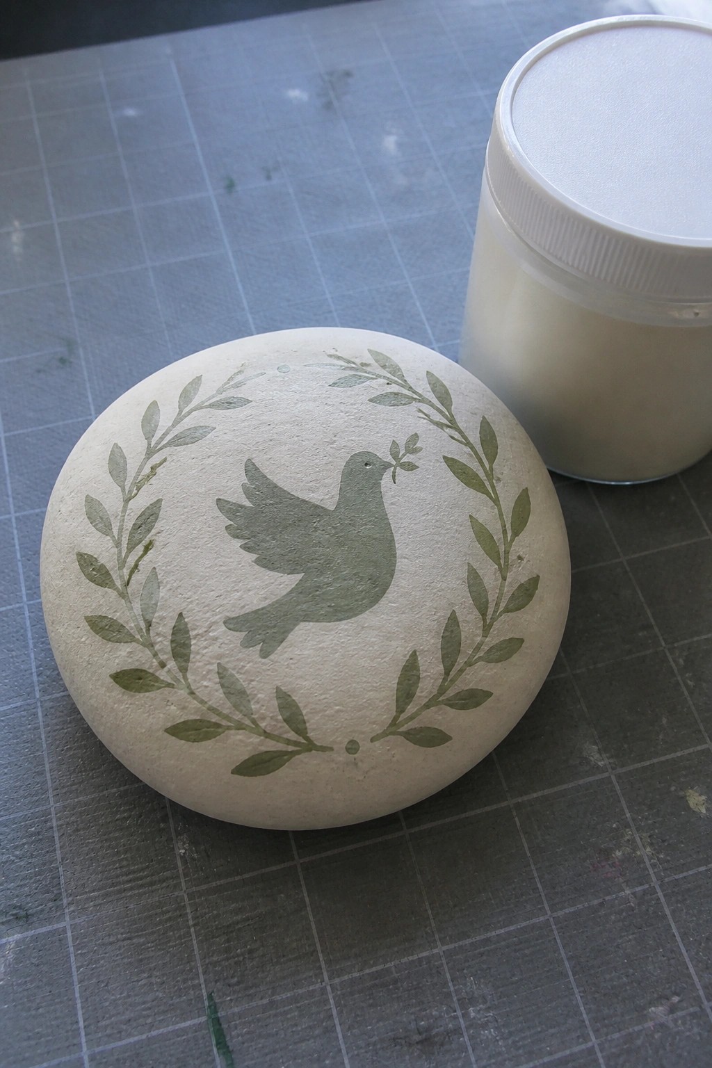 Round stone painted with green dove and leafy wreath on gray grid mat