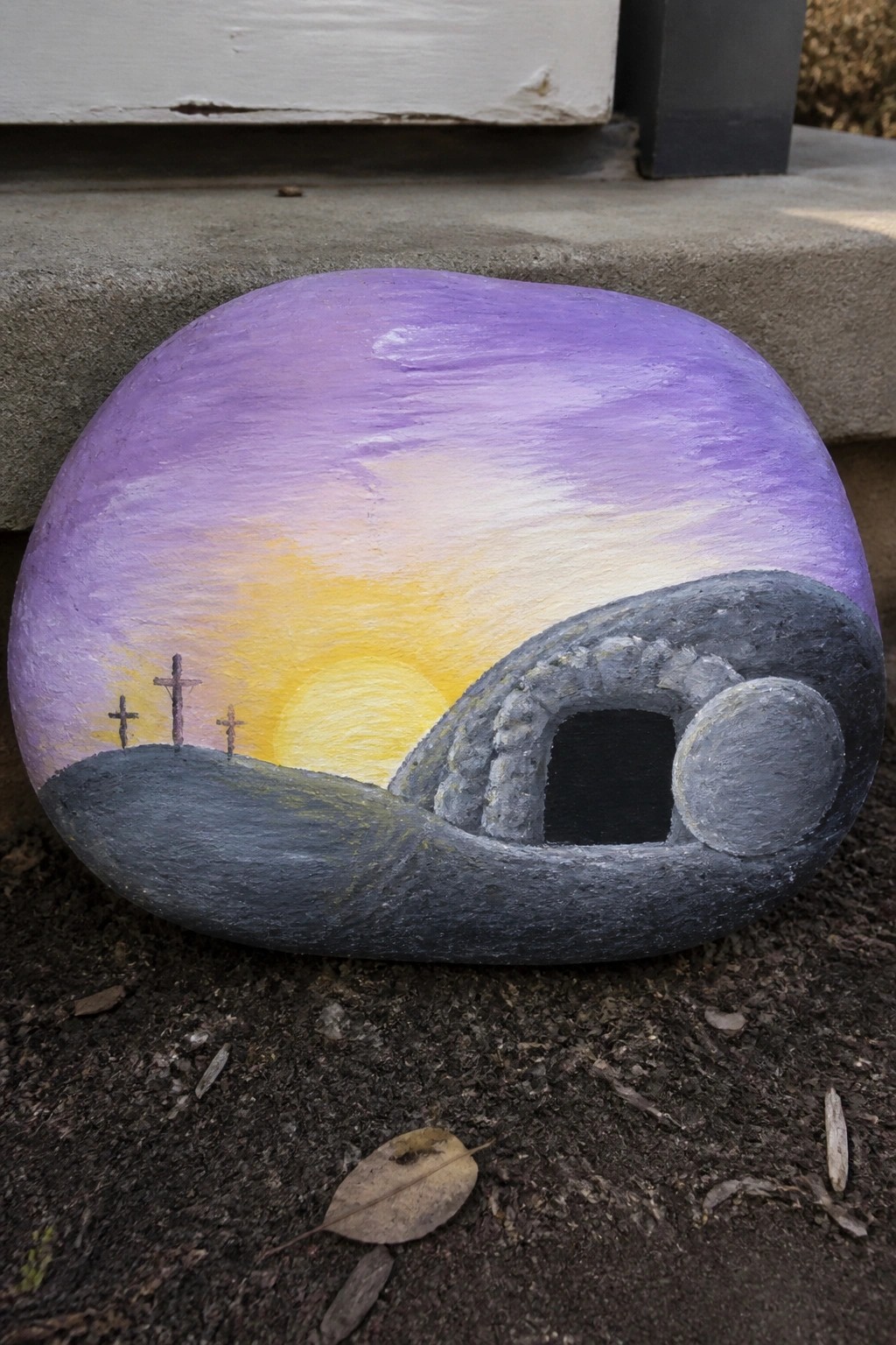 Painted rock showing purple sunset, three crosses, and stone tomb with rolled-away boulder.