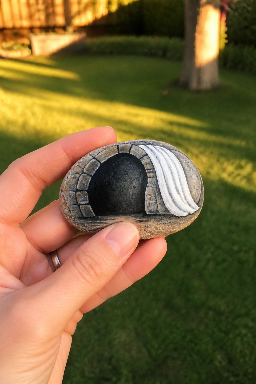 A hand holds a painted oval rock showing a dark arched tomb entrance with gray stone details and white lines on one side.