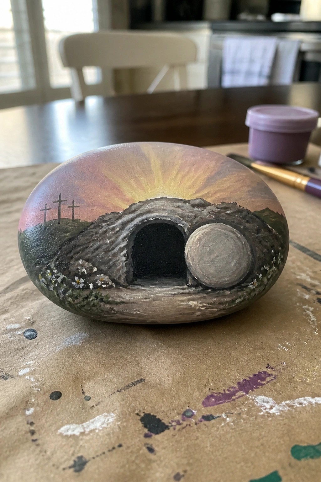 Painted rock shows sunrise over hill with three crosses and empty tomb with stone