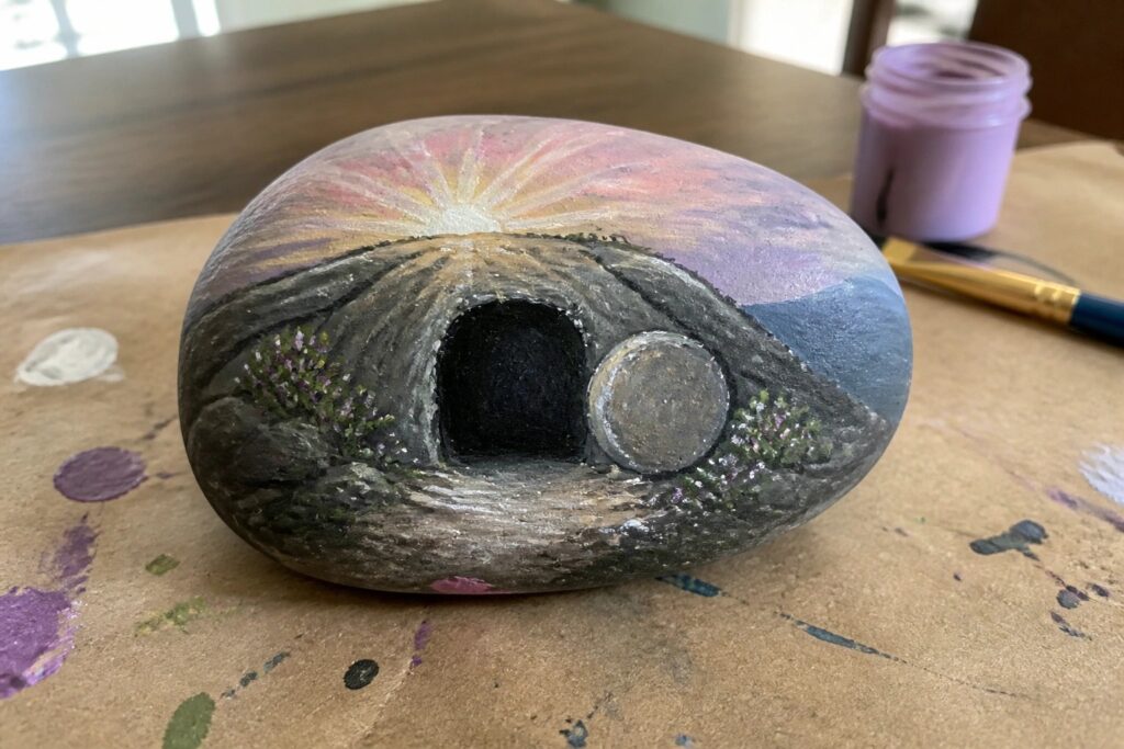 Painted rock shows sunrise over hill with three crosses and empty tomb with stone