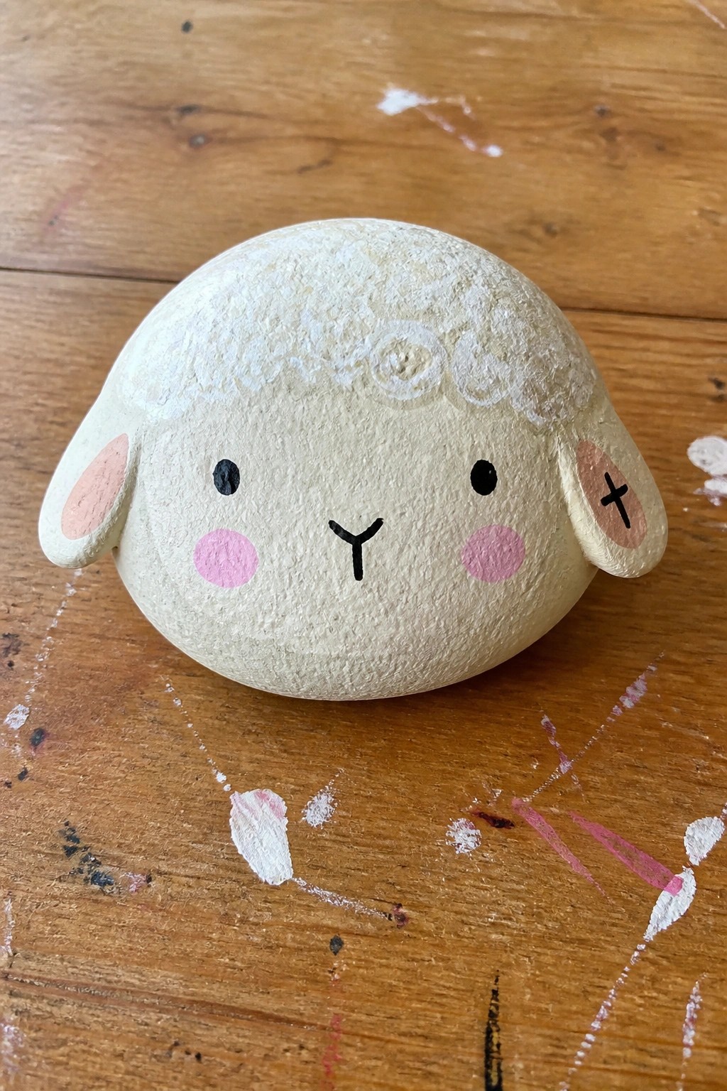 Round off-white ceramic sheep figurine with pink cheeks and ears on paint-stained wood