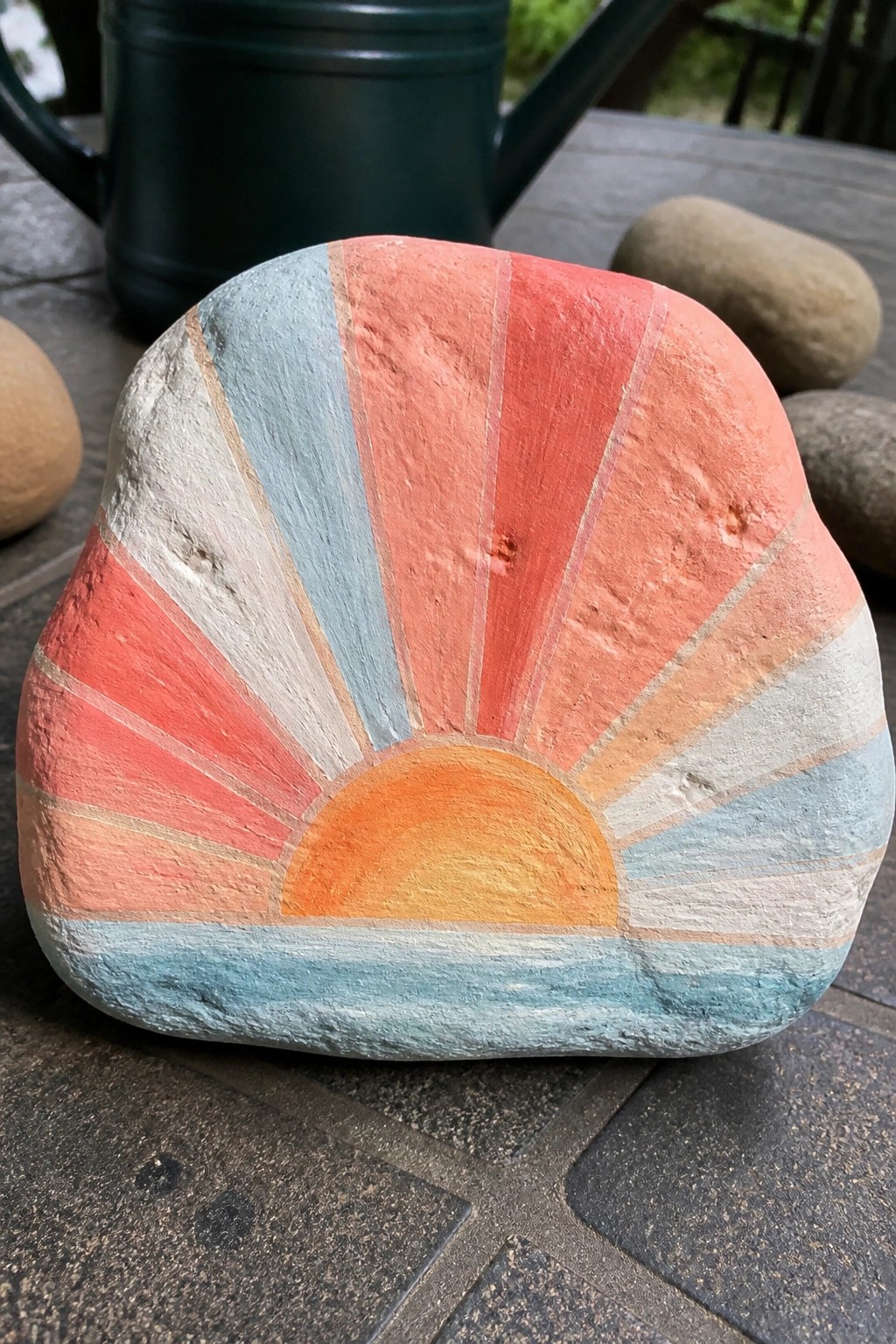Painted rock with vibrant sunset: orange sun, pink-orange rays, and blue waves.