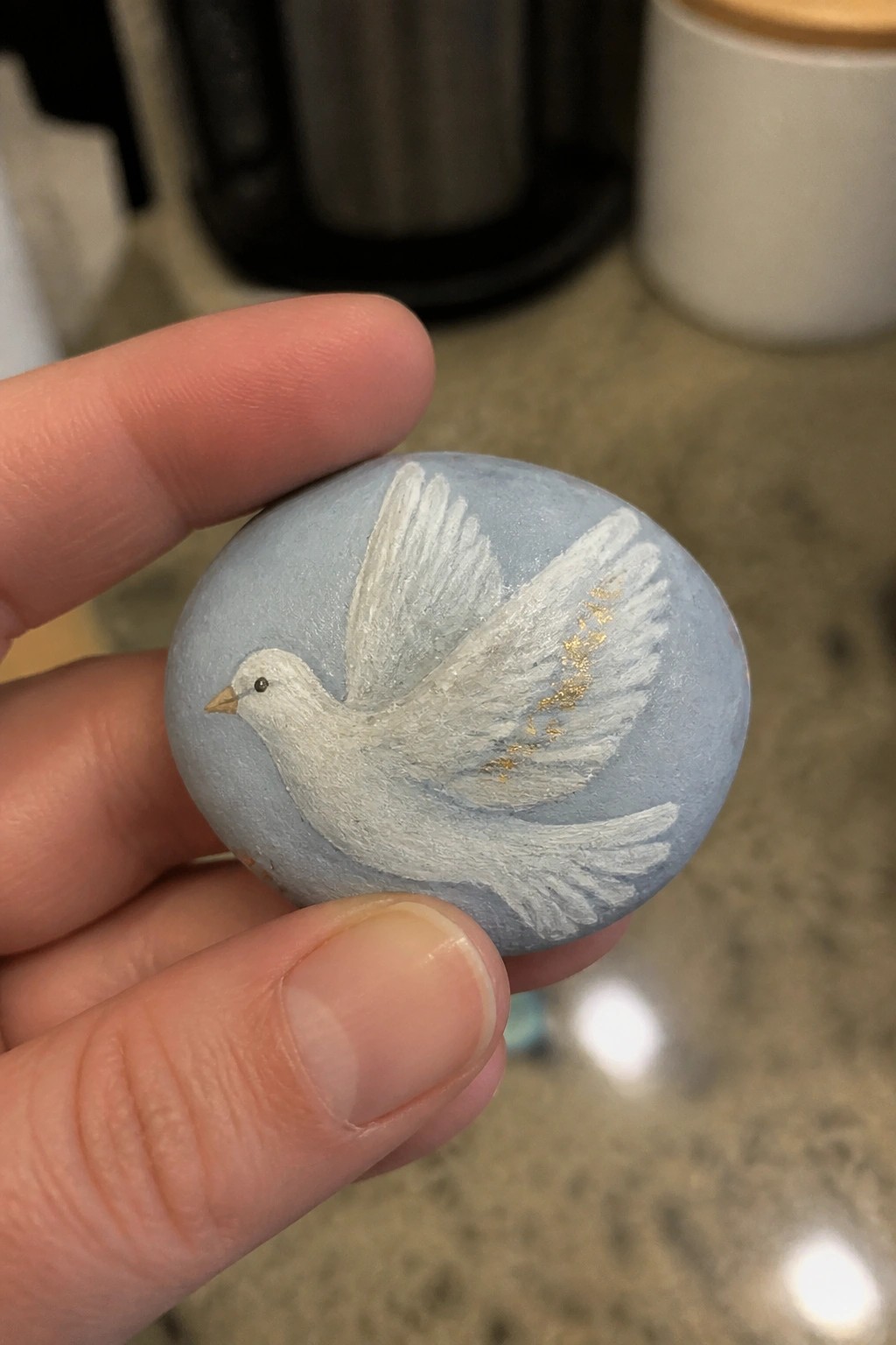 Hand holding light blue rock painted with white dove and gold wing accents