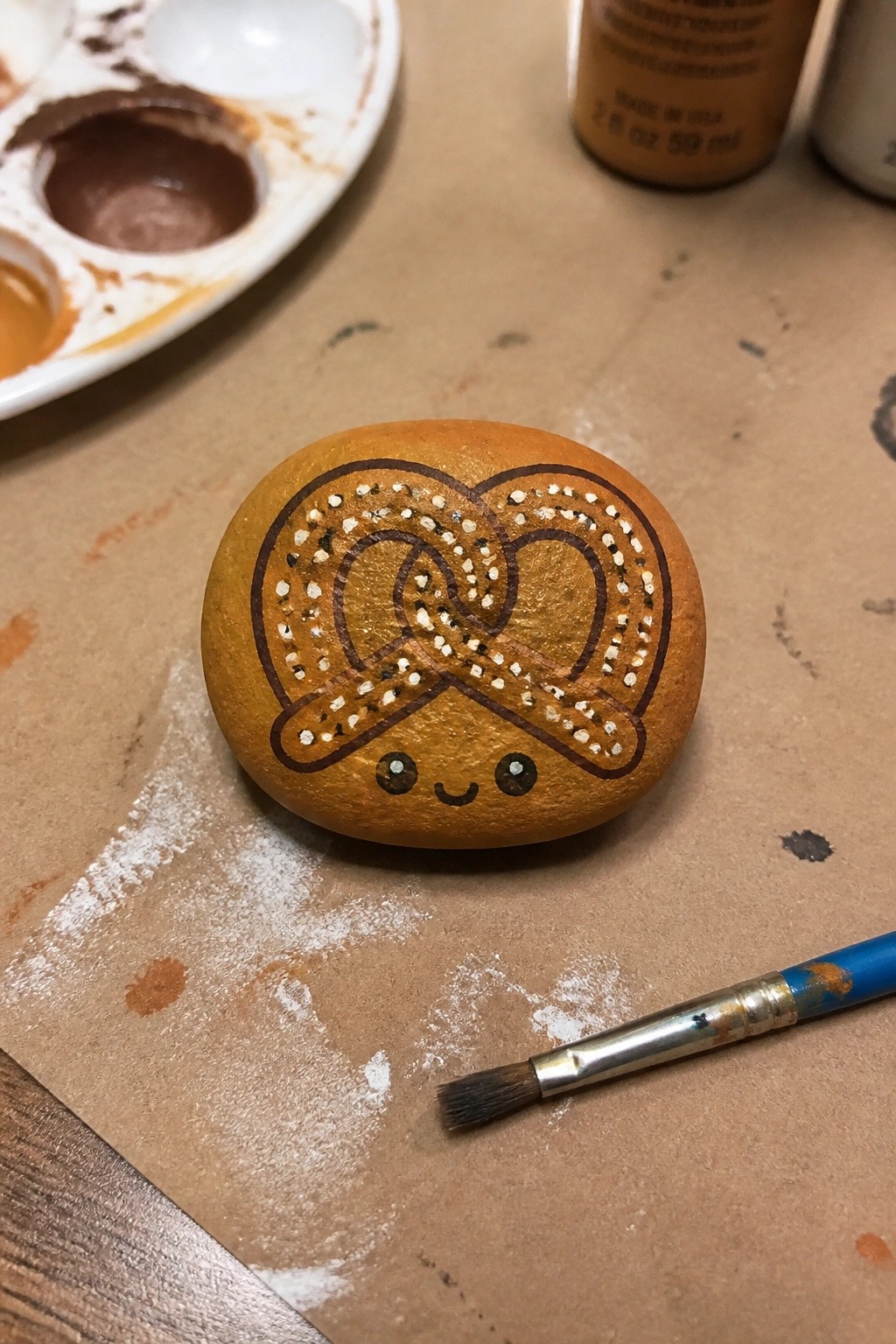 A painted brown rock shaped like a pretzel with white dots and a cute face sits on brown paper next to a paintbrush and palette.